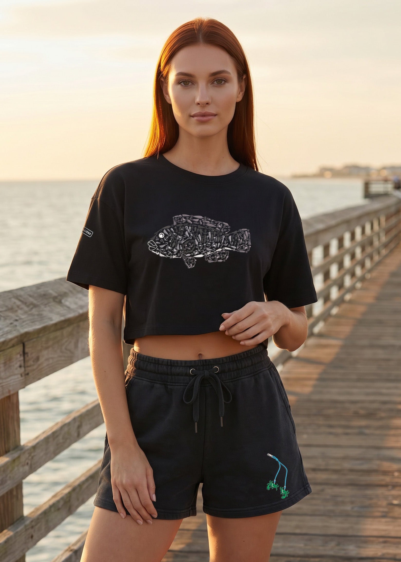 Woman wearing a black t-shirt and shorts with a fish design on a wooden pier by the water.