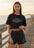 Woman wearing a black t-shirt and shorts with a fish design on a wooden pier by the water.