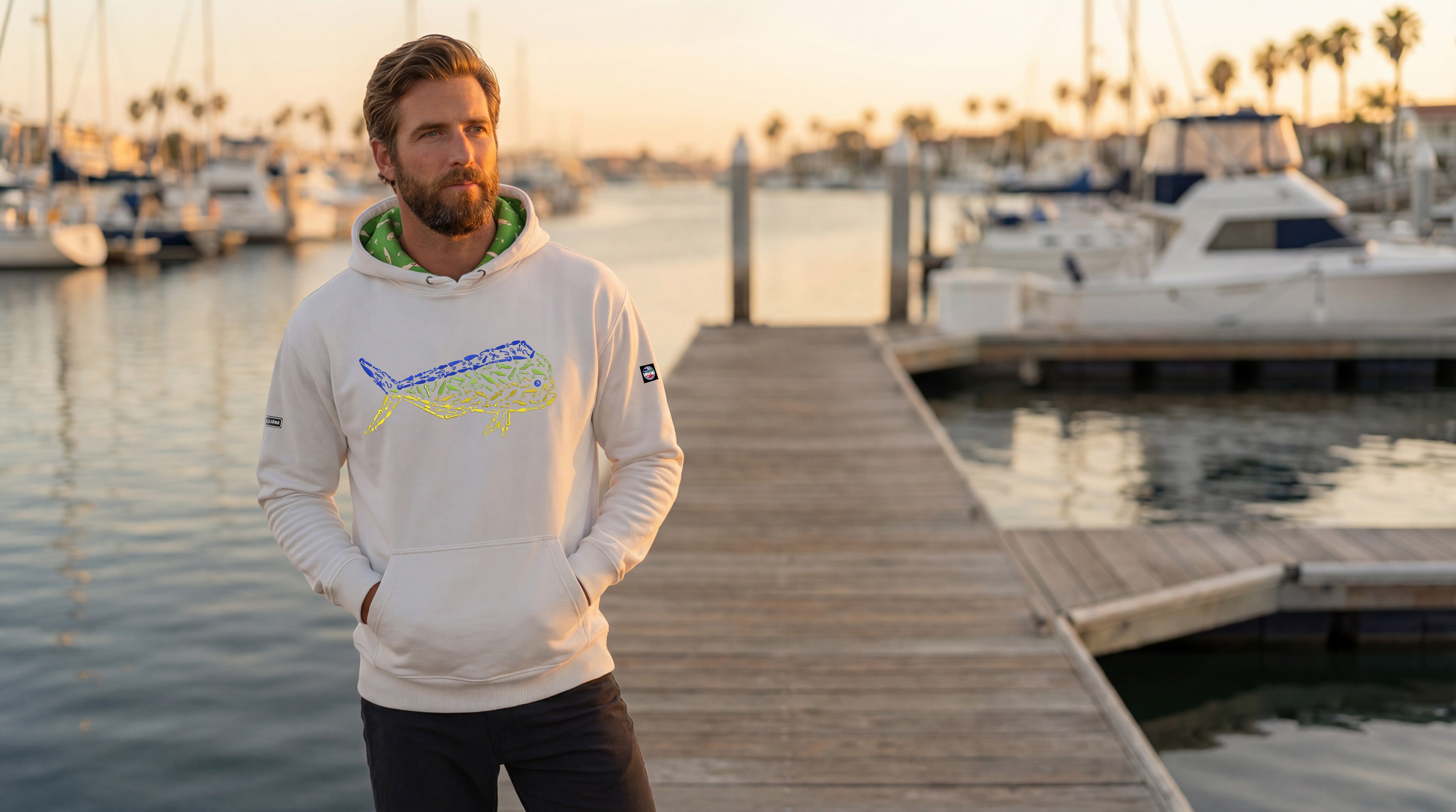 Man wearing a hoodie with a logo on a dock with boats in the background