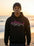 Person wearing a black hoodie with a pink and white shark design on a beach at sunset.