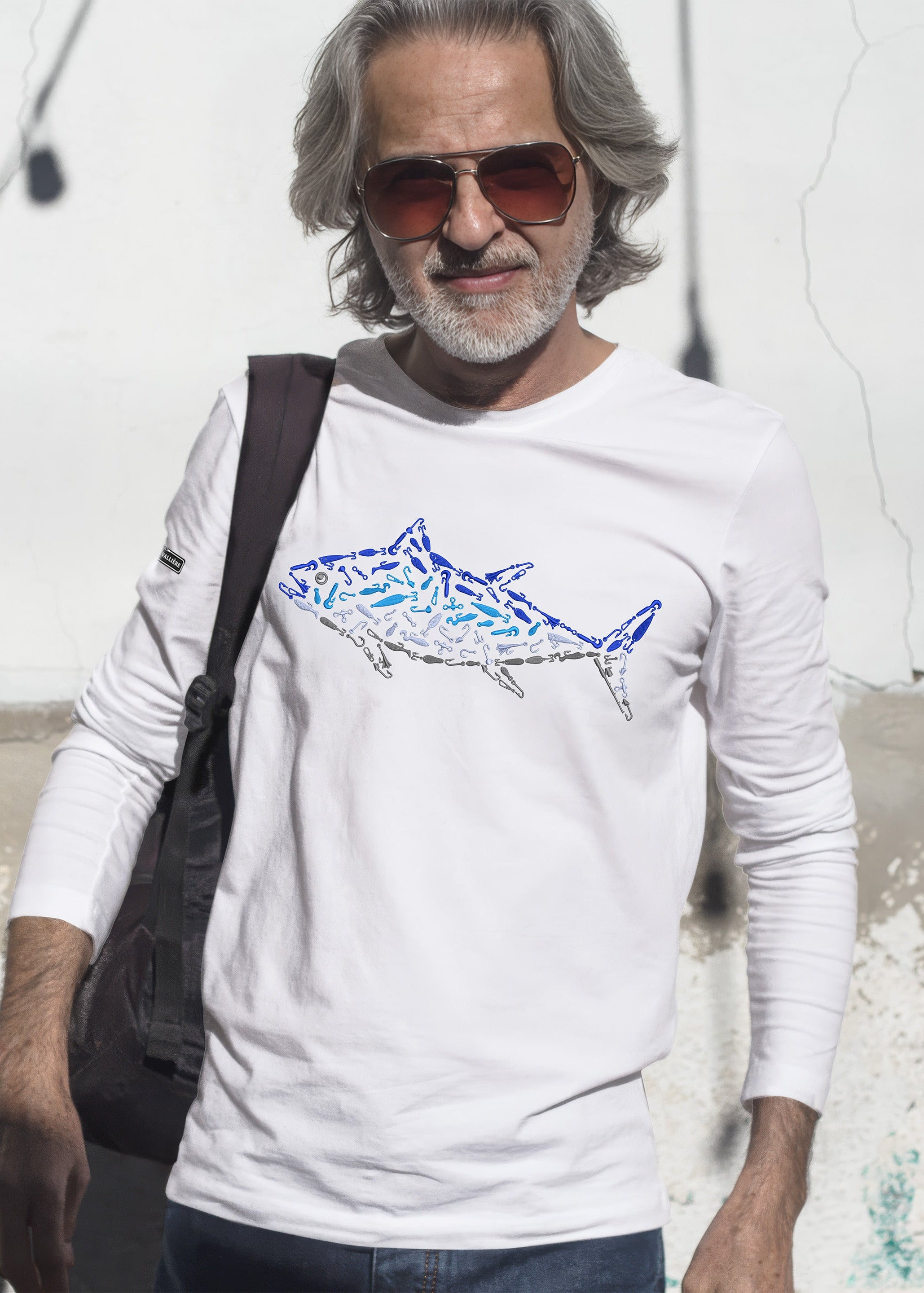 Man wearing a white t-shirt with a shark design, sunglasses, and a black bag against a white wall.