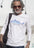 Man wearing a white t-shirt with a shark design, sunglasses, and a black bag against a white wall.