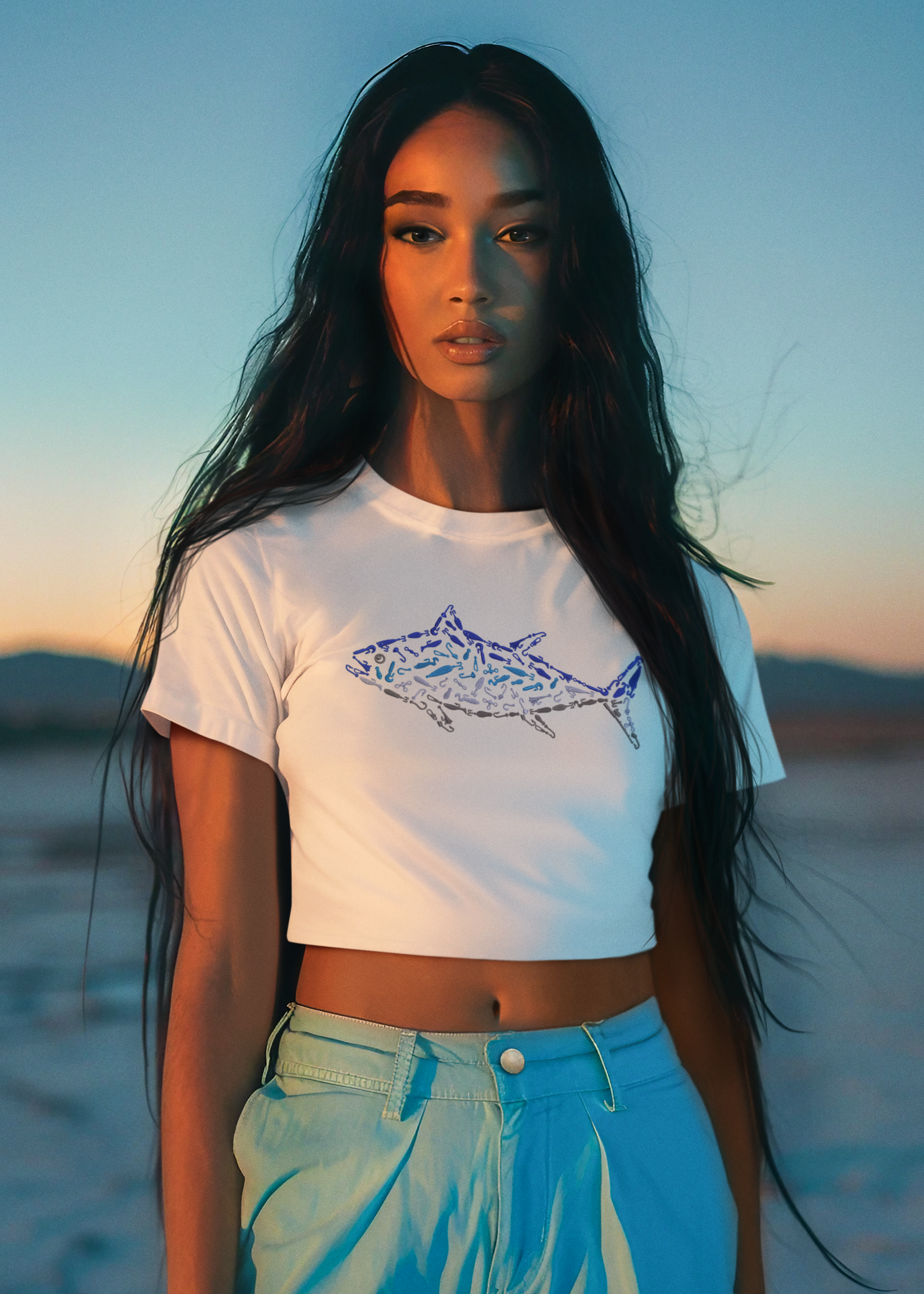 A woman wearing a white crop tee with a graphic print of a tuna fish, paired with blue shorts, standing at a beach during sunset.